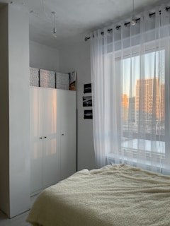 A minimalist bedroom features a cozy bed with a cream-colored fluffy blanket. To the side, there is a large white wardrobe with storage boxes on top. Sheer white curtains cover a window, allowing soft natural light to filter in. The room's color palette is neutral, and the view outside shows buildings reflecting the warm glow of sunset.