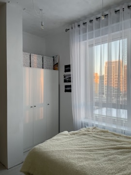 A minimalist bedroom features a cozy bed with a cream-colored fluffy blanket. To the side, there is a large white wardrobe with storage boxes on top. Sheer white curtains cover a window, allowing soft natural light to filter in. The room's color palette is neutral, and the view outside shows buildings reflecting the warm glow of sunset.