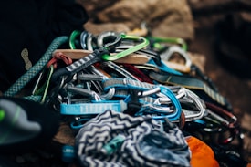 A collection of colorful climbing gear including carabiners and straps is piled together. The image shows a variety of equipment with different colors and materials, suggesting preparedness for rock climbing.