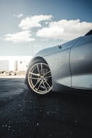 A close-up view of the front side of a car, focusing on a polished alloy wheel with a reflective silver body. The background features a semi-truck with a trailer parked on a concrete surface. The sky is partly cloudy with a bright, daytime atmosphere.