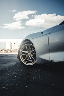 A close-up view of the front side of a car, focusing on a polished alloy wheel with a reflective silver body. The background features a semi-truck with a trailer parked on a concrete surface. The sky is partly cloudy with a bright, daytime atmosphere.