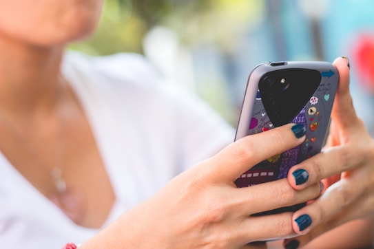 A person with blue painted nails is holding a smartphone with a colorful and decorated case. The focus is on the hands and phone, with a blurred background suggesting an outdoor setting.