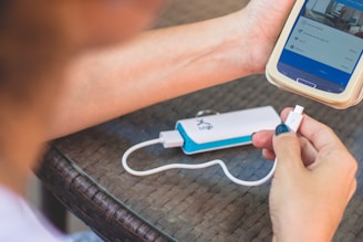 A person holding a smartphone with one hand and a USB cable with the other. The phone is connected to a white and blue portable charger that is resting on a woven surface.