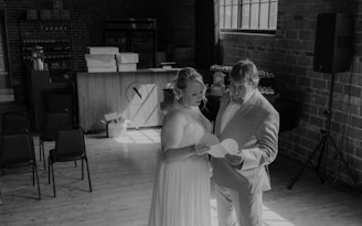 A couple dressed in formal attire stands in a dimly lit room with exposed brick walls. They are holding and looking at a piece of paper together. Several empty chairs, a bar counter, and decorations, including stacked boxes and small items, can be seen in the background. Sunlight streams in through a large window, casting shadows across the wooden floor.