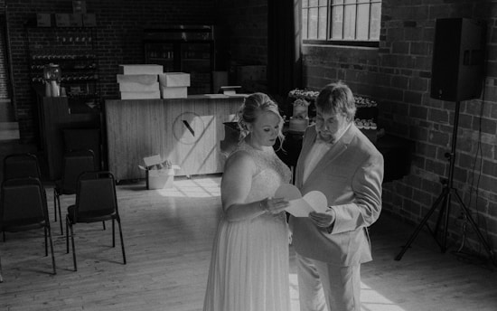A couple dressed in formal attire stands in a dimly lit room with exposed brick walls. They are holding and looking at a piece of paper together. Several empty chairs, a bar counter, and decorations, including stacked boxes and small items, can be seen in the background. Sunlight streams in through a large window, casting shadows across the wooden floor.