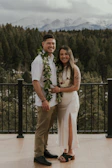 A couple stands on a wooden deck with a forest and snow-capped mountains in the background. Both are smiling and dressed in formal attire. The man wears a white shirt with a leafy garland, while the woman is in a white dress with a floral lei. The setting appears peaceful and natural.