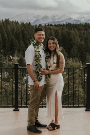 A couple stands on a wooden deck with a forest and snow-capped mountains in the background. Both are smiling and dressed in formal attire. The man wears a white shirt with a leafy garland, while the woman is in a white dress with a floral lei. The setting appears peaceful and natural.