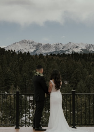 A couple stands on a balcony overlooking a forest with snow-capped mountains in the background. The woman is in a white dress and the man is in a dark suit with a floral boutonniere. They appear to be in a serene setting with overcast skies.