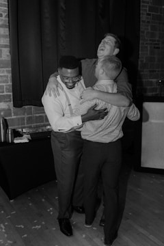 Three people are embracing and laughing together in a candid moment, showcasing joy and camaraderie. They are standing on a wooden floor with a curtain and brick wall in the background.