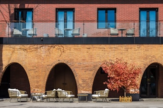 A brick building features both upper and lower levels. The upper level includes a balcony with a glass railing and several wicker chairs and tables. The lower level displays a series of archways with stylish outdoor furniture placed beneath them, including cushioned chairs and a small table. A vibrant red-leaved tree stands to the right, adding a splash of color against the brick facade.