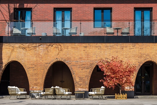 A brick building features both upper and lower levels. The upper level includes a balcony with a glass railing and several wicker chairs and tables. The lower level displays a series of archways with stylish outdoor furniture placed beneath them, including cushioned chairs and a small table. A vibrant red-leaved tree stands to the right, adding a splash of color against the brick facade.