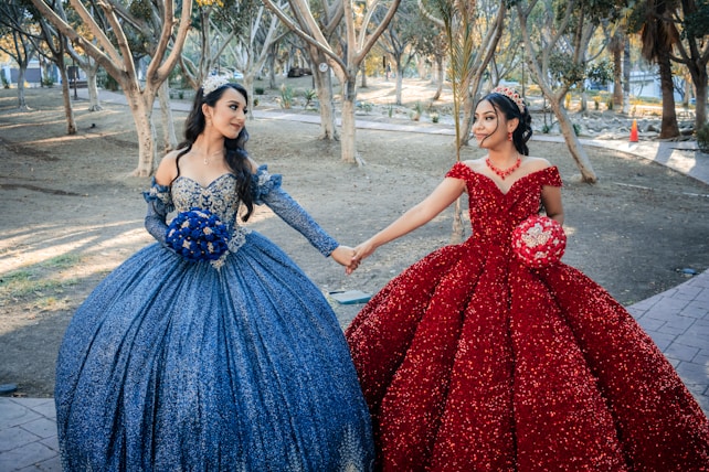 Two women in elaborate ball gowns holding hands in a park setting. One woman wears a sparkling blue dress with a matching bouquet, while the other is in a shimmering red dress and holds a red bouquet. Both are wearing tiaras and appear to be enjoying a festive occasion.