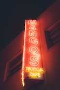 A neon sign for Mango's Tropical Cafe illuminated in vibrant red and yellow colors against a dark background, attached to the side of a building.