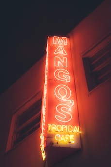 A neon sign for Mango's Tropical Cafe illuminated in vibrant red and yellow colors against a dark background, attached to the side of a building.
