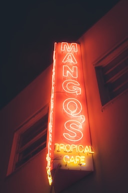 A neon sign for Mango's Tropical Cafe illuminated in vibrant red and yellow colors against a dark background, attached to the side of a building.