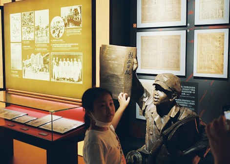 A girl stands next to a museum exhibit featuring a bronze statue of a person holding a large book. Behind them, illuminated displays showcase historical photographs and documents on the wall.