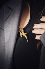 A close-up of a person wearing a dark ribbed shirt partially covered by a gray jacket. A large gold-colored brooch, shaped like a stylized insect, is pinned to the shirt. The person's fingernails, painted black, rest gently on the garment.