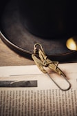 A detailed, brass-colored mechanical grasshopper-like paperclip lies on a newspaper. The background features a dark ceramic cup and saucer, with a small metallic spoon partially visible. The overall aesthetic is vintage and artisanal.