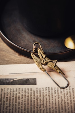A detailed, brass-colored mechanical grasshopper-like paperclip lies on a newspaper. The background features a dark ceramic cup and saucer, with a small metallic spoon partially visible. The overall aesthetic is vintage and artisanal.