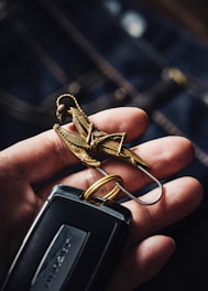 A hand is holding a set of keys. The focus is on a detailed metal keychain shaped like a mechanical insect or grasshopper. The keychain is intricately designed with metallic accents. The keys attached include a modern car key with a sleek black design.