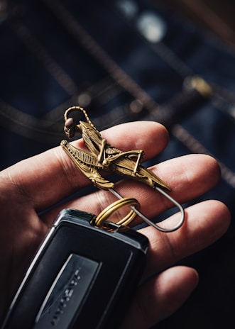 A hand is holding a set of keys. The focus is on a detailed metal keychain shaped like a mechanical insect or grasshopper. The keychain is intricately designed with metallic accents. The keys attached include a modern car key with a sleek black design.