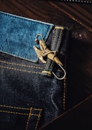 A close-up view of a dark blue denim fabric with contrasting yellow stitching. Attached to the denim is a metallic keychain or accessory that resembles a bird or an intricate design. The texture of the denim and the sheen of the metal create a rich contrast.