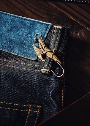 A close-up view of a dark blue denim fabric with contrasting yellow stitching. Attached to the denim is a metallic keychain or accessory that resembles a bird or an intricate design. The texture of the denim and the sheen of the metal create a rich contrast.
