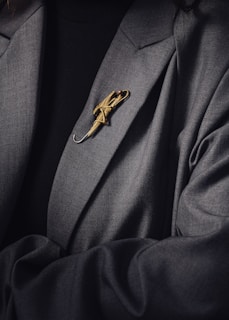 A person is wearing a dark suit jacket adorned with a decorative gold brooch shaped like an umbrella. The brooch is intricately designed and stands out against the dark fabric of the clothing.
