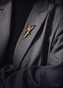 A person is wearing a dark suit jacket adorned with a decorative gold brooch shaped like an umbrella. The brooch is intricately designed and stands out against the dark fabric of the clothing.