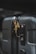 A close-up of a dark-colored suitcase featuring a gold bird-shaped ornament attached to the zipper. The suitcase has a textured surface with visible zippers and a TSA-approved lock. The lighting highlights the ornately designed bird accessory, contrasting against the suitcase's smooth, dark body.