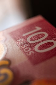 A close-up view of a currency bill displaying the number 100 and the word 'PESOS' in bold white text against a textured red background. The image is slightly blurred around the edges, giving focus to the central text. The design includes intricate patterns and a subtle gradient of colors.