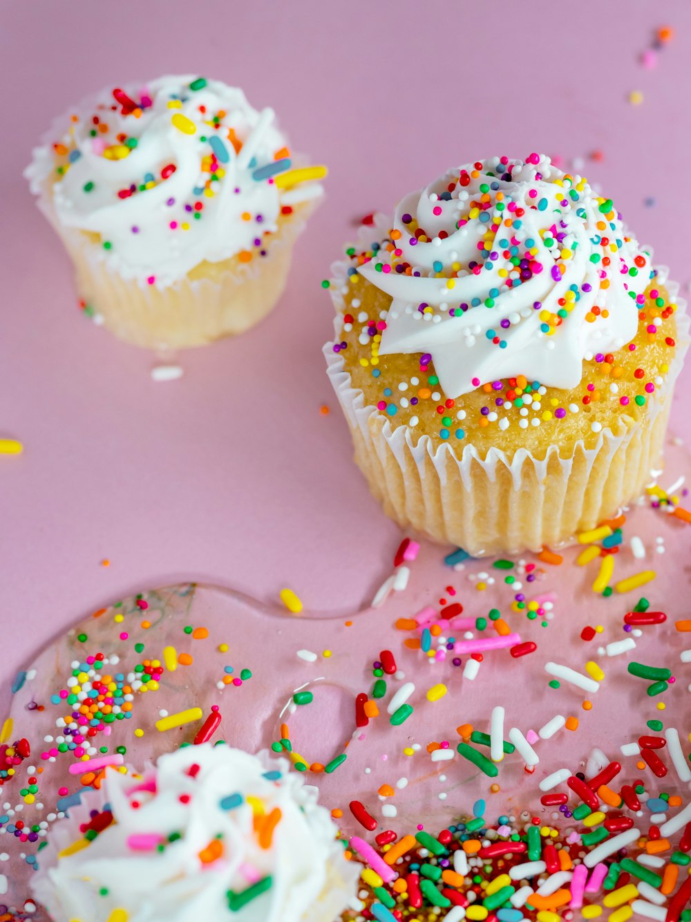 three cupcakes with white frosting and sprinkles on a pink