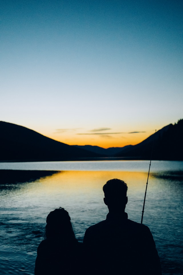 Two friends laughing together while fishing on a calm lake at sunrise.
