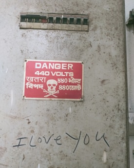 A weathered and rusty metal electrical box featuring a red warning sign that reads 'Danger 440 Volts' along with multilingual text, including Hindi. A small skull and crossbones icon is present beneath the text. There are several black switches above the sign, and graffiti below it reads 'I love you' in black marker.