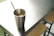 A stainless steel cup filled with iced coffee and a black straw sits on a shiny metal table. The table is positioned next to wooden stools, and the lighting creates soft reflections on the surfaces.