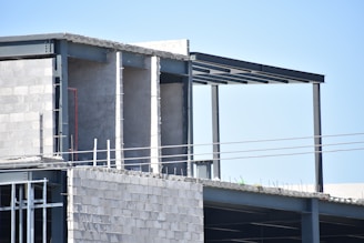 An unfinished building under construction with visible concrete blocks and steel beams. The structure appears to be at an early stage with some parts exposed and not yet completed. There are electrical conduits and reinforcement rods visible.