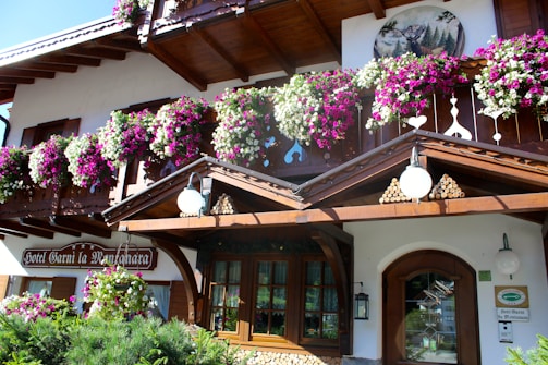 A charming wooden chalet-style building adorned with vibrant pink, purple, and white flowers. The structure features intricate wooden carvings and a picturesque painting of a deer in a forest. A neatly arranged pile of logs and traditional lanterns enhance the rustic aesthetic.