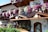 A charming wooden chalet-style building adorned with vibrant pink, purple, and white flowers. The structure features intricate wooden carvings and a picturesque painting of a deer in a forest. A neatly arranged pile of logs and traditional lanterns enhance the rustic aesthetic.