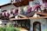 A charming wooden chalet-style building adorned with vibrant pink, purple, and white flowers. The structure features intricate wooden carvings and a picturesque painting of a deer in a forest. A neatly arranged pile of logs and traditional lanterns enhance the rustic aesthetic.