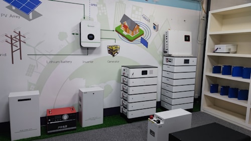 The image depicts a showroom or demonstration area showcasing various energy storage and conversion devices. Several white battery systems are stacked or aligned neatly. In the background, there is a diagram on the wall illustrating a solar energy system, including components like PV arrays, lithium batteries, inverters, and generators. A few blue battery cells are displayed on shelves to the right.