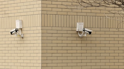 Two surveillance cameras are mounted on a corner of a beige brick building. The cameras are positioned at a right angle to each other and are installed on a cream-colored brick wall. A barren tree branch is visible on the right side.