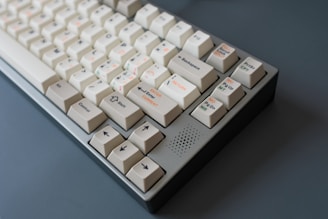 A mechanical keyboard with off-white keycaps and multicolored legends. The keyboard features several keys with symbols and text, including arrow keys and function keys. The case is grey, and the overall design has a vintage or retro feel.