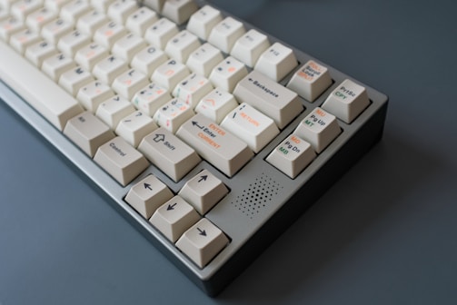 A mechanical keyboard with off-white keycaps and multicolored legends. The keyboard features several keys with symbols and text, including arrow keys and function keys. The case is grey, and the overall design has a vintage or retro feel.
