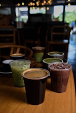 A selection of six drinks in various cups is arranged on a wooden table inside a dimly lit cafe. The drinks include iced green tea, a chocolate beverage with ice, and a black cup containing coffee with a crema layer. The background features blurred lighting and chairs, contributing to a cozy atmosphere.