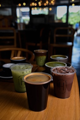 A selection of six drinks in various cups is arranged on a wooden table inside a dimly lit cafe. The drinks include iced green tea, a chocolate beverage with ice, and a black cup containing coffee with a crema layer. The background features blurred lighting and chairs, contributing to a cozy atmosphere.