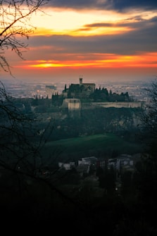 A picturesque landscape with an ancient castle perched on a hill, surrounded by lush greenery and silhouetted against a dramatic sunset sky. The vibrant hues of orange and yellow in the sky contrast with the dark silhouette of the trees and the castle.