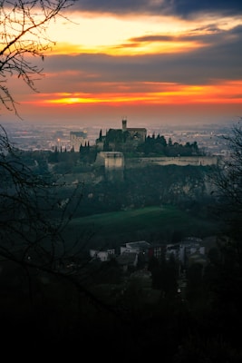 A picturesque landscape with an ancient castle perched on a hill, surrounded by lush greenery and silhouetted against a dramatic sunset sky. The vibrant hues of orange and yellow in the sky contrast with the dark silhouette of the trees and the castle.