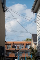 A residential area with a clear blue sky as the backdrop. The scene is framed by two buildings on each side with a row of apartment buildings in the background. Several telecommunication wires crisscross the upper portion of the frame. Clothes are hanging outside on the balconies of the apartments, drying in the open air.