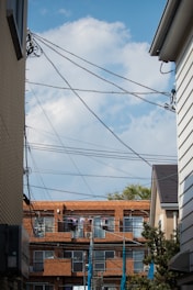 A residential area with a clear blue sky as the backdrop. The scene is framed by two buildings on each side with a row of apartment buildings in the background. Several telecommunication wires crisscross the upper portion of the frame. Clothes are hanging outside on the balconies of the apartments, drying in the open air.