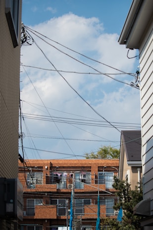 A residential area with a clear blue sky as the backdrop. The scene is framed by two buildings on each side with a row of apartment buildings in the background. Several telecommunication wires crisscross the upper portion of the frame. Clothes are hanging outside on the balconies of the apartments, drying in the open air.